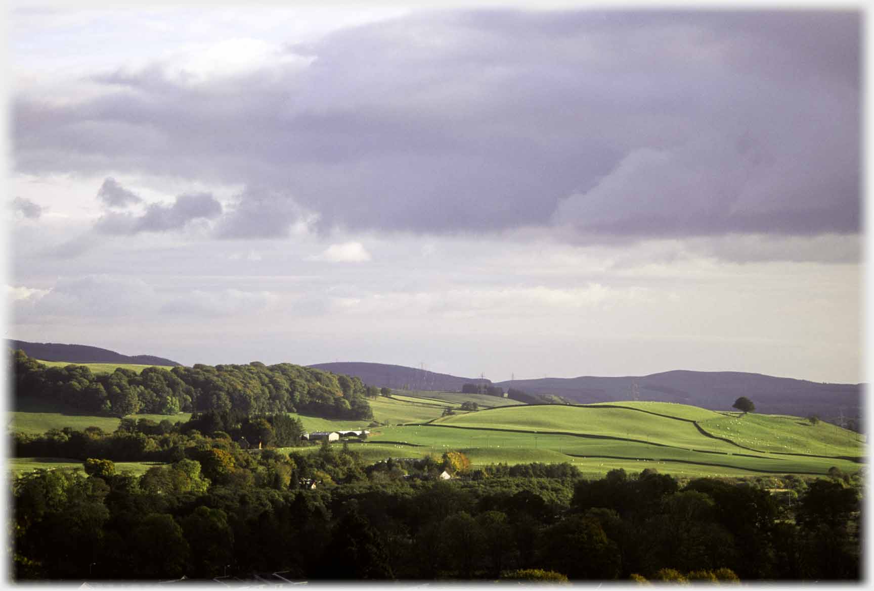 Fields to right, trees to left and foreground, dark cloud over.