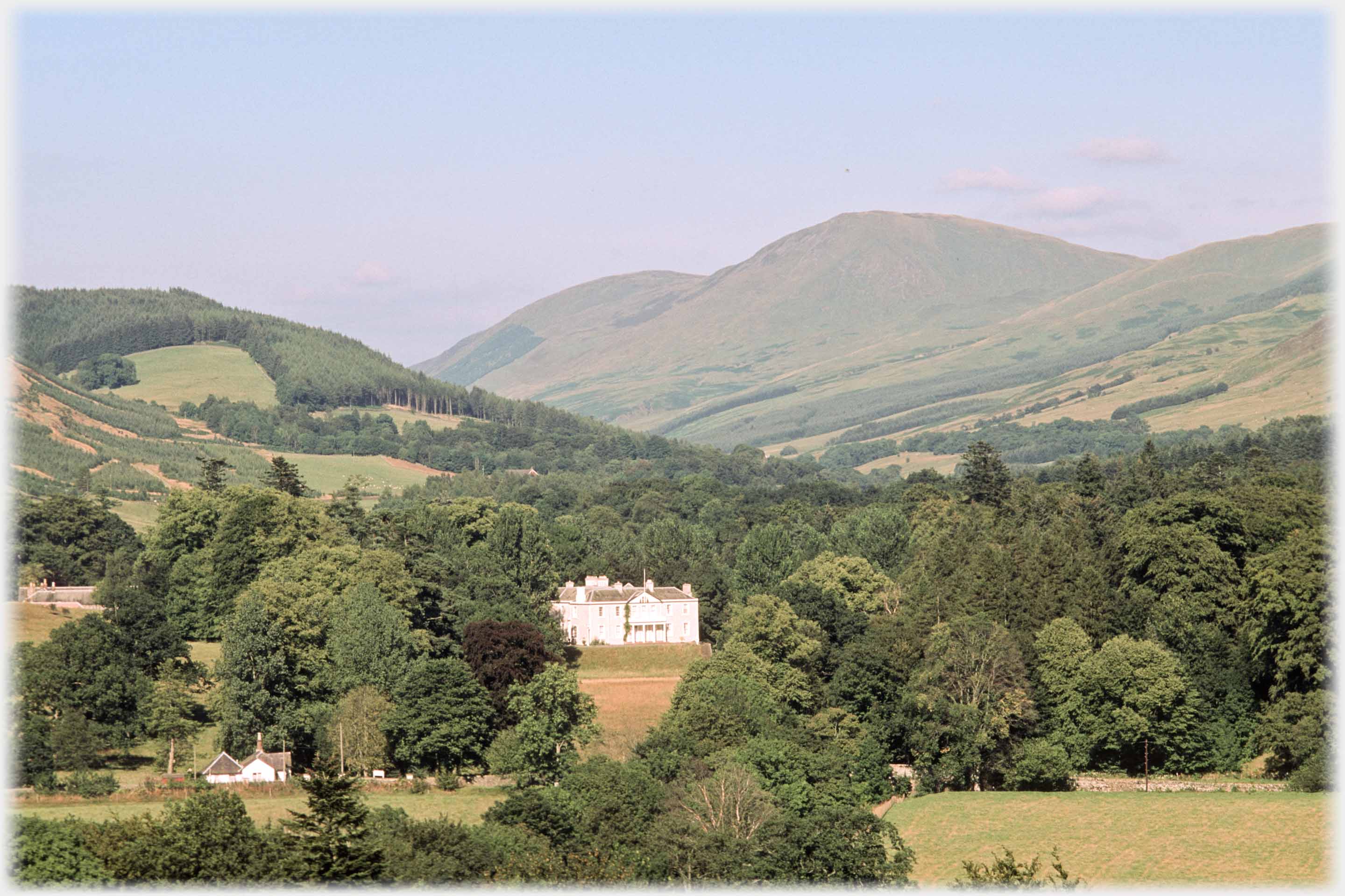 Mansion house set in  among large trees, hills beyond.