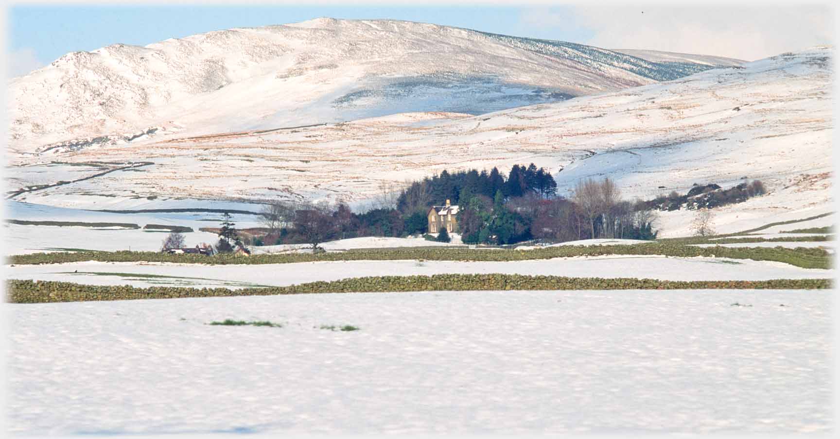 Snow covered field and landscape, house in clump of trees, hill as background.