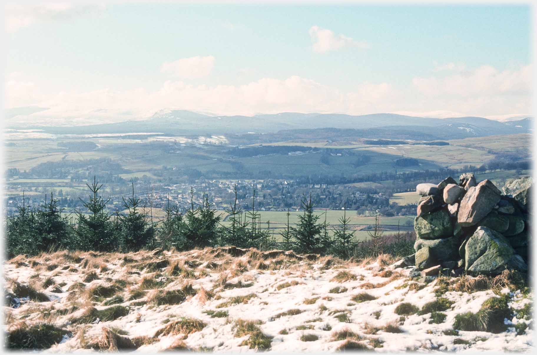 Light snow covering over grassy topped hill with cairn and twon in distance.