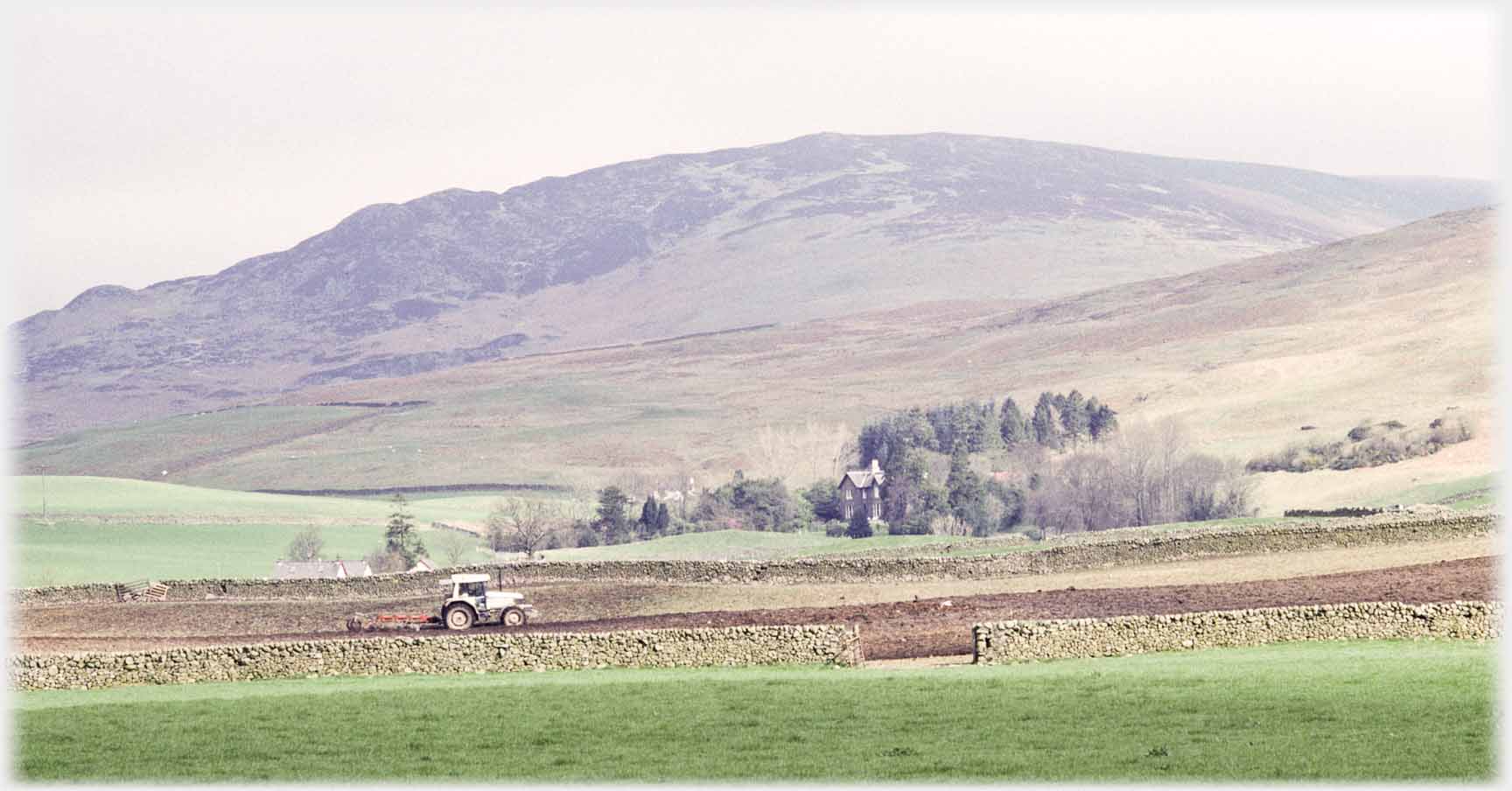 Field being ploughed by tractor, house in clump of trees, hill as background.