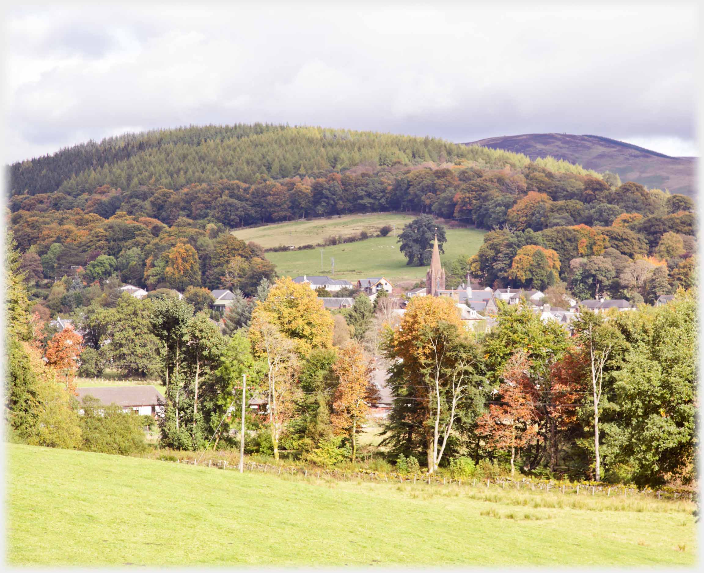 Autumn turning trees in foreground houses behind, autumnal trees on hill beyond.
