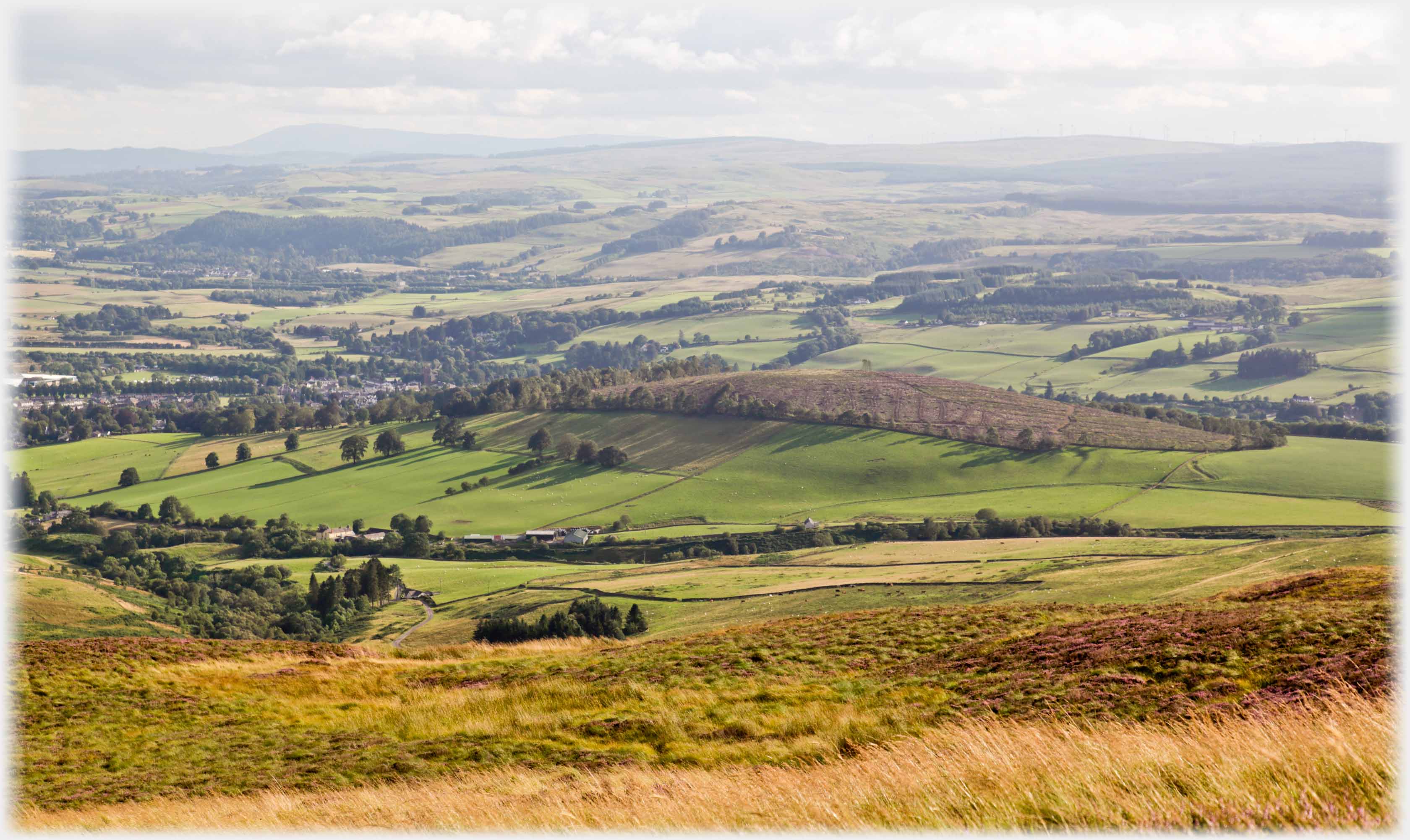 Trees and houses to left, grassed hill to right, fields in foreground.