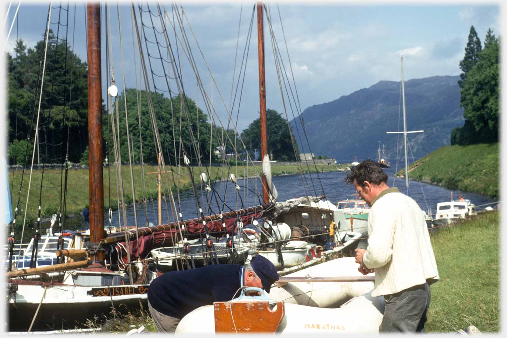 Two men working on inflatable dinghy, yachts and canal behind them.