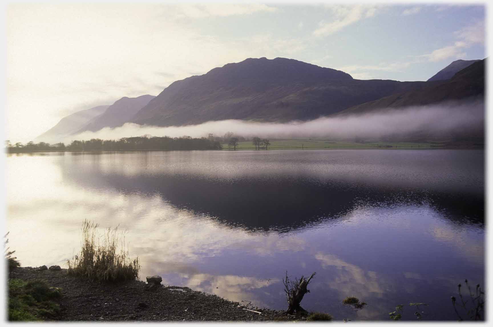 Looking across loch with still water, waterside of fields with layer of mist low on them, hill above.