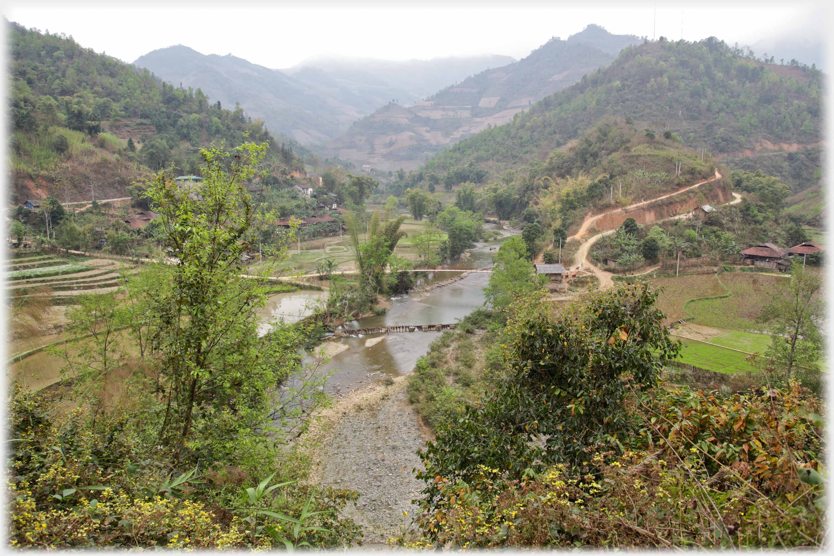 River widing between hills with footbridge, barrages and paths.