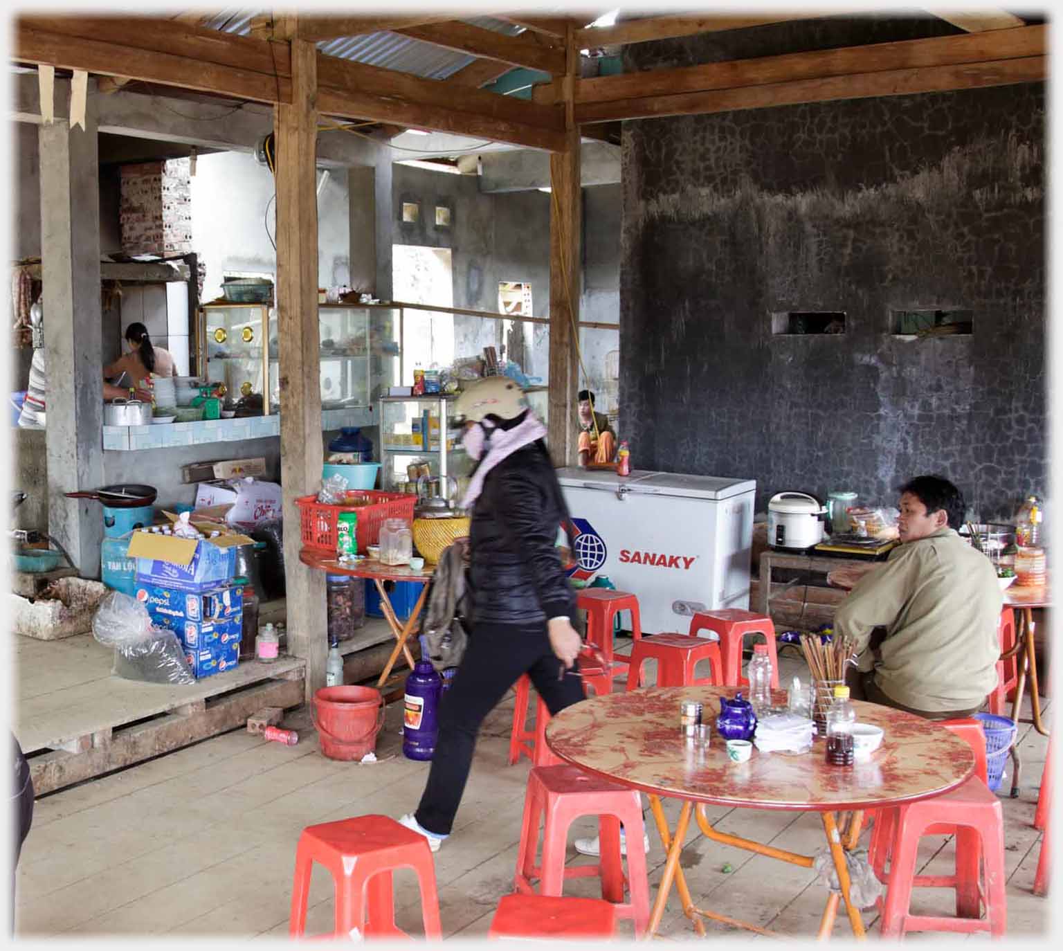 Corner of cafe with walking customer, sitting customer and discernable kitchen work in background.