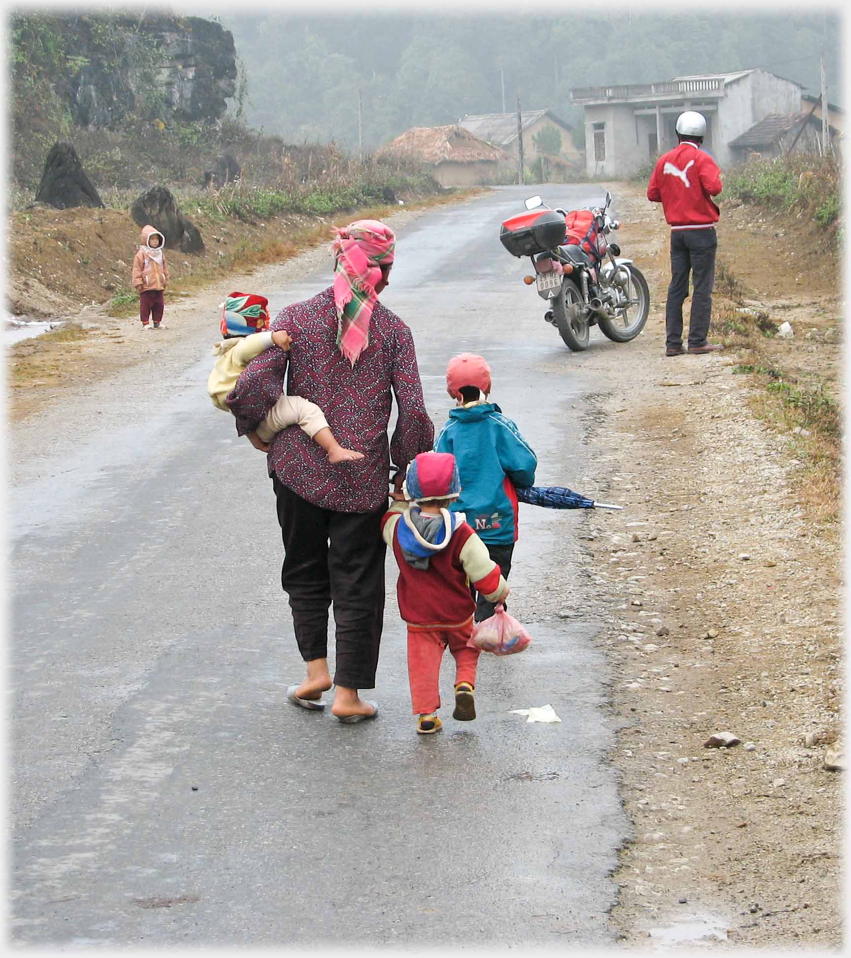 Woman one child on hip, leading two others, one with umbrella, another child watching man by motorbike.