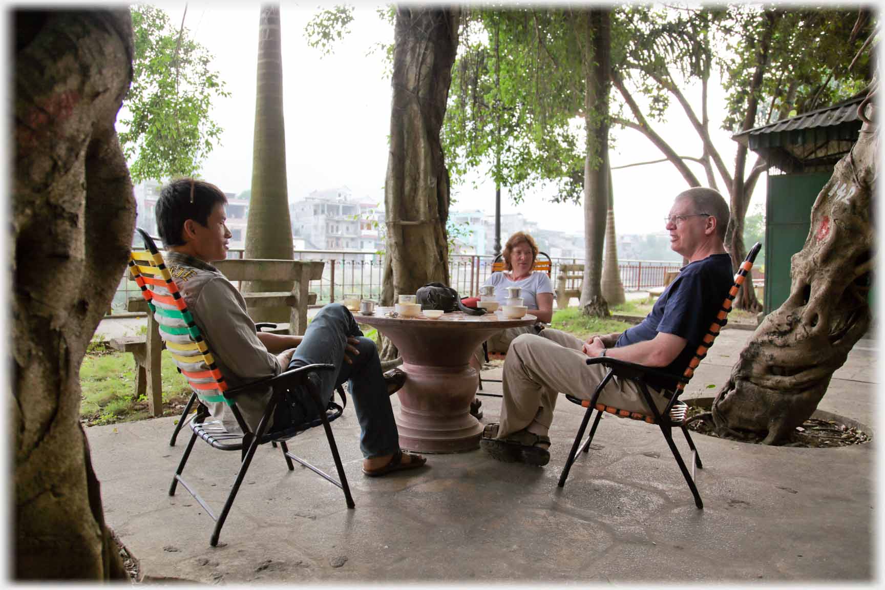 Three people sitting round table in treed open courtyard.