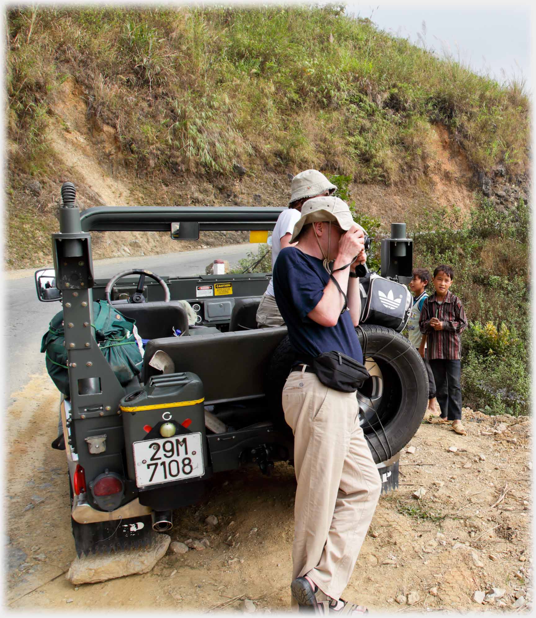 Man leaning on Jeep taking photo, two boys near-by.
