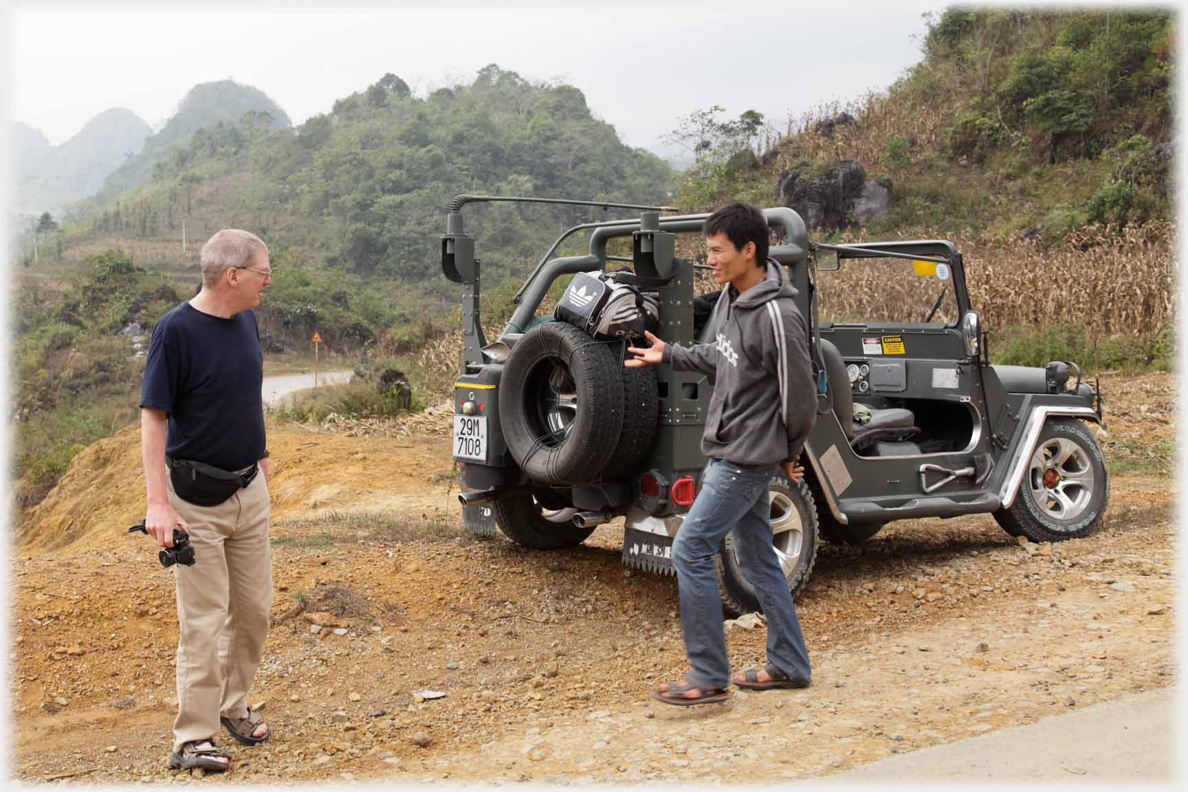 Two men by open Jeep in country, one holding hand out and open.