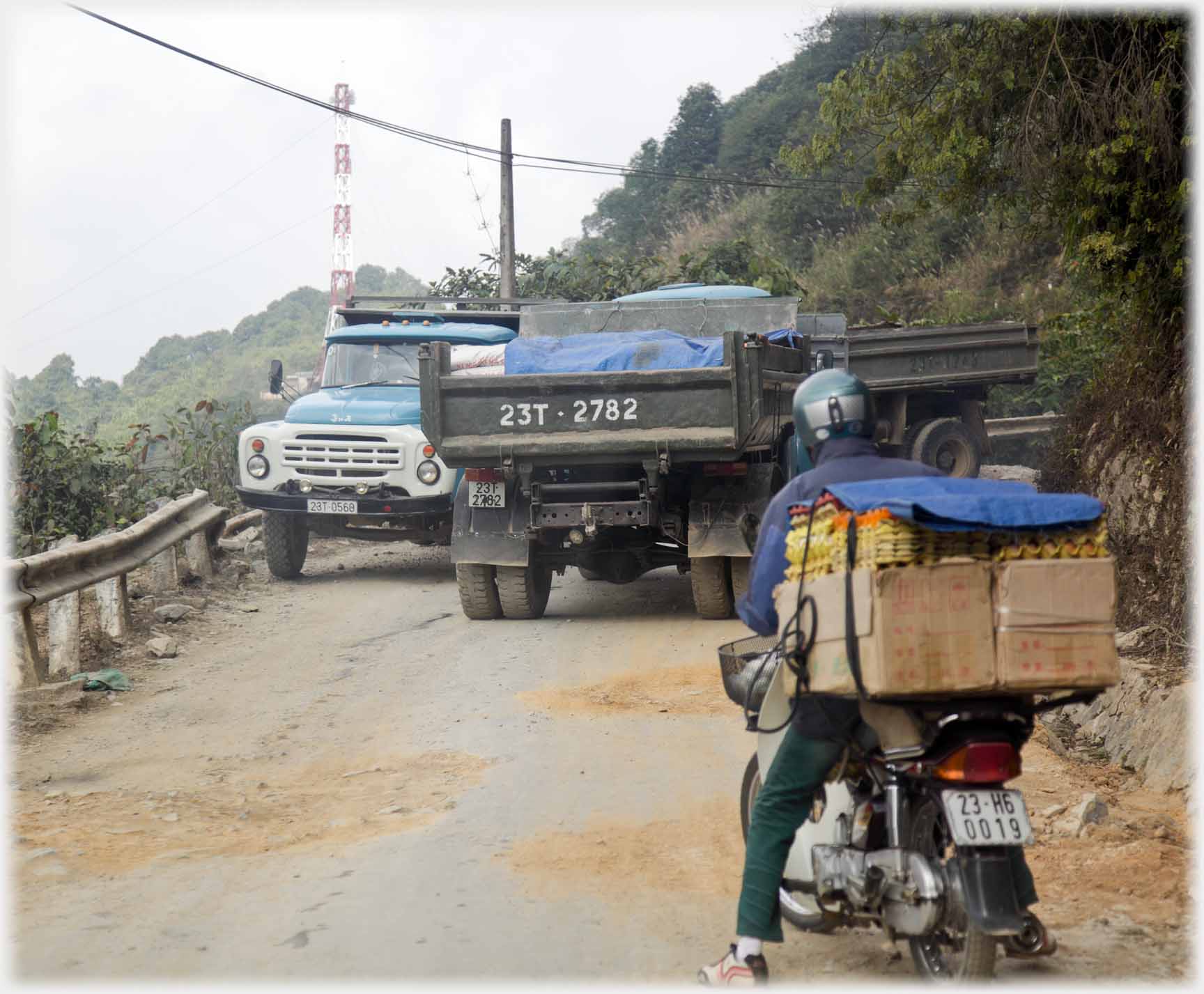Two lorries struggling to pass one another while motorcyclist with many egg boxes on back waits.