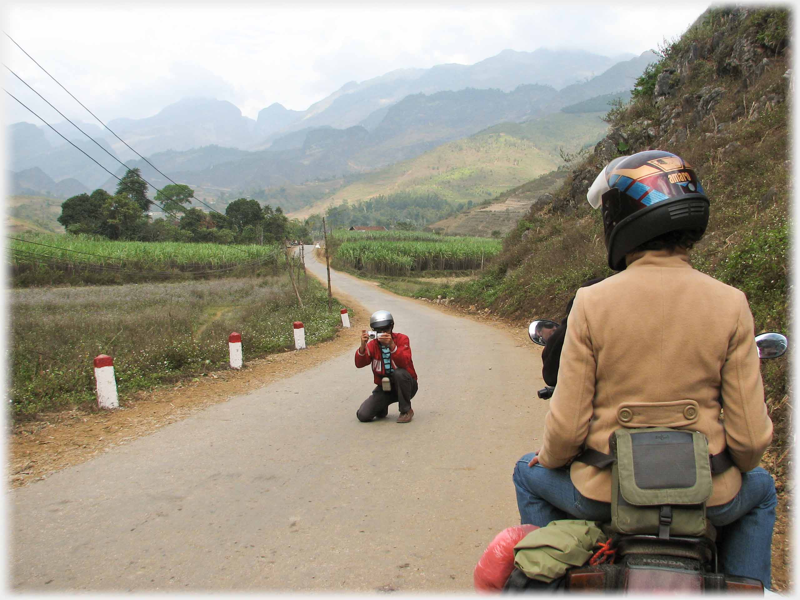 Han croaching in middle of the road taking photo of couple on motorbike.