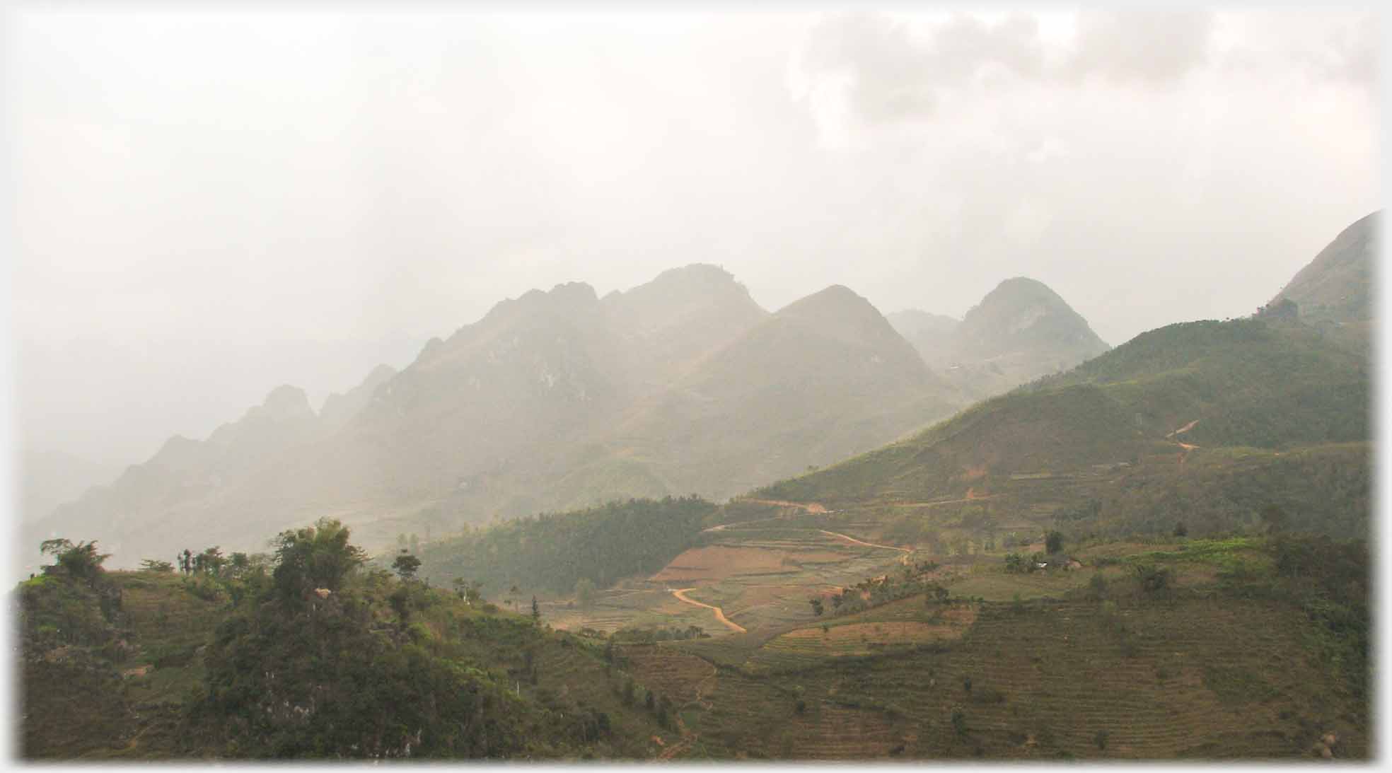 Misty peaks behind misty country with tracks, roads, and trees.