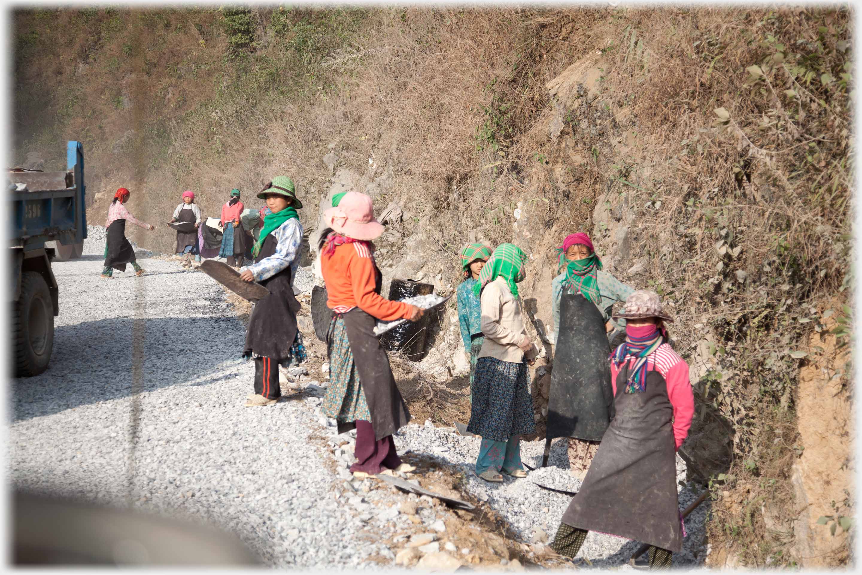 A dozen or so women, in black aprons, some holding containers of stones on newly laid road and ditch.