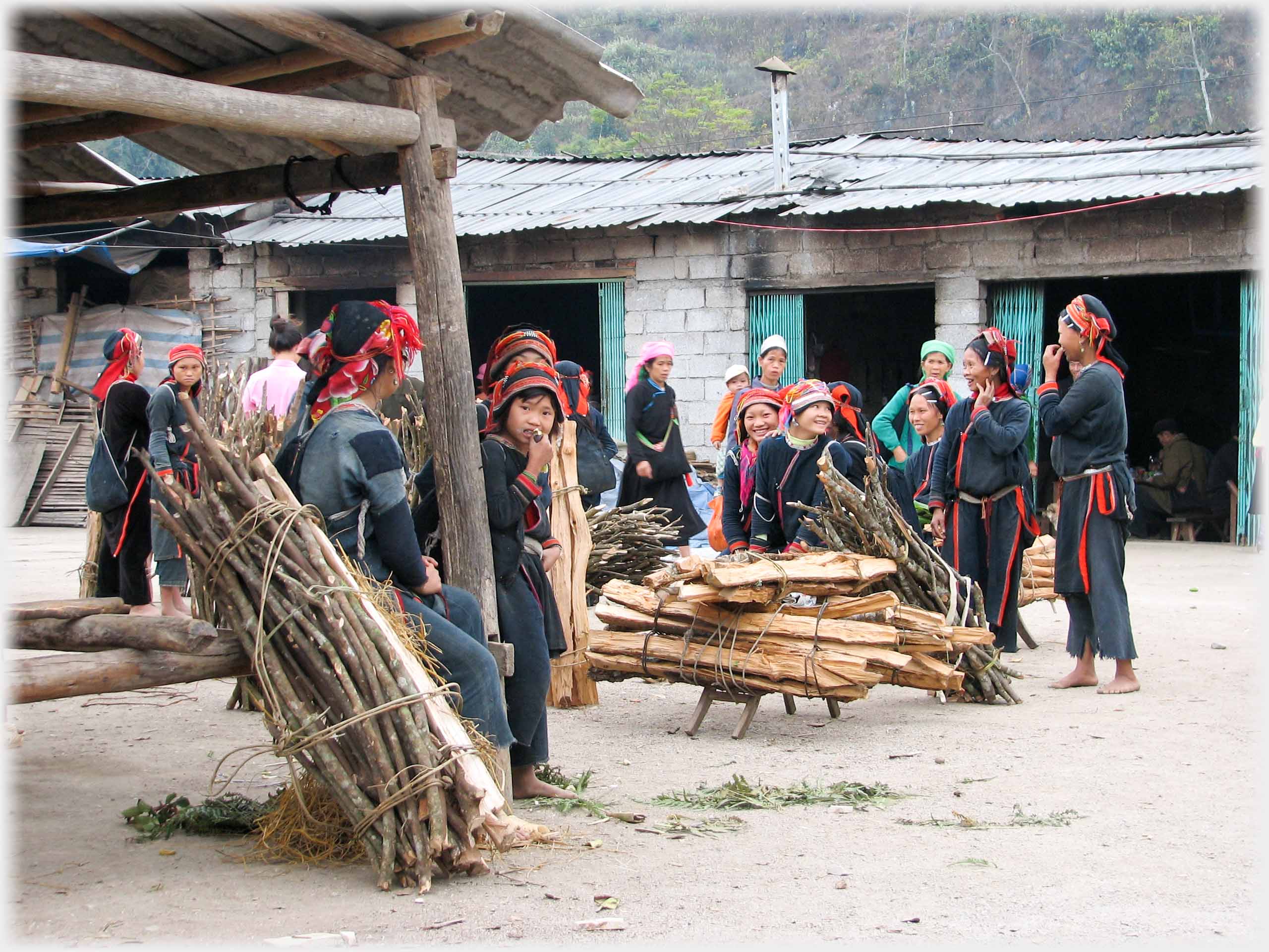 Group of women in yard wearing black and red clothes with bundles of wood.