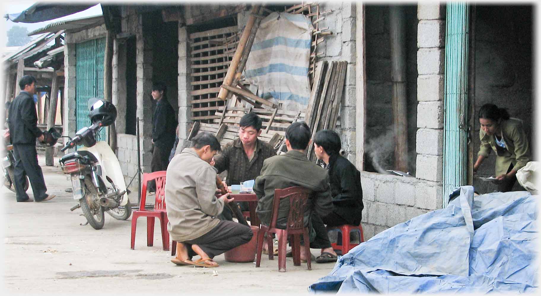 Four men at low table by rough tarpaulins.
