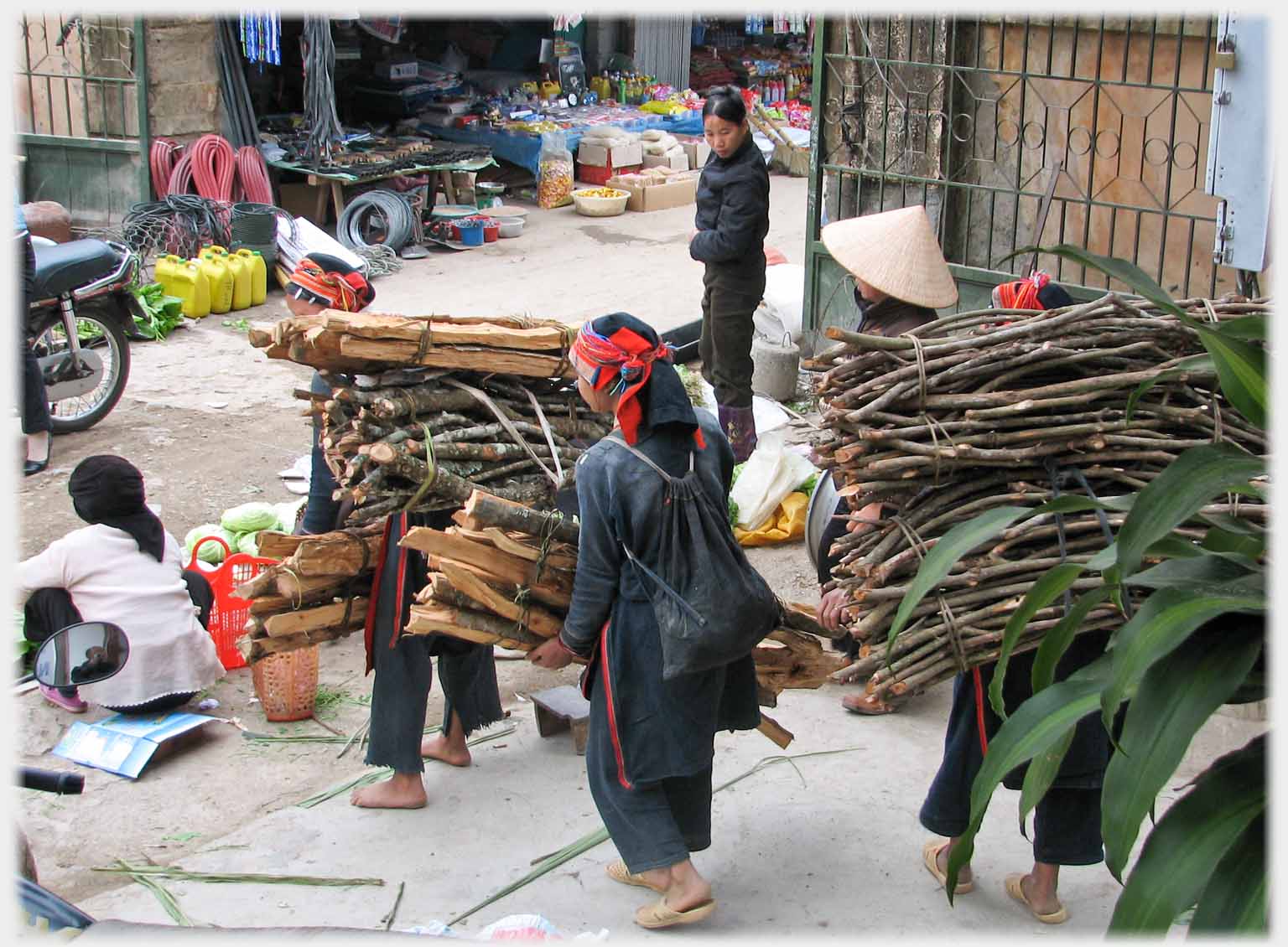 Three women carrying bundles of wood walking out into street.