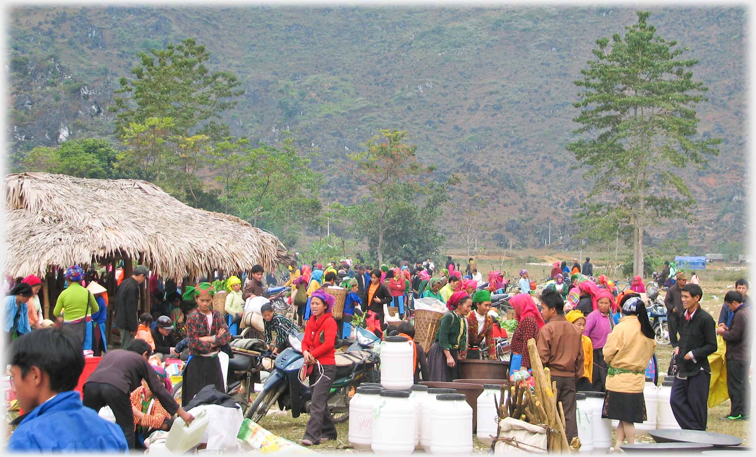 Crowds with goods for sale, thatched hut, background hills.
