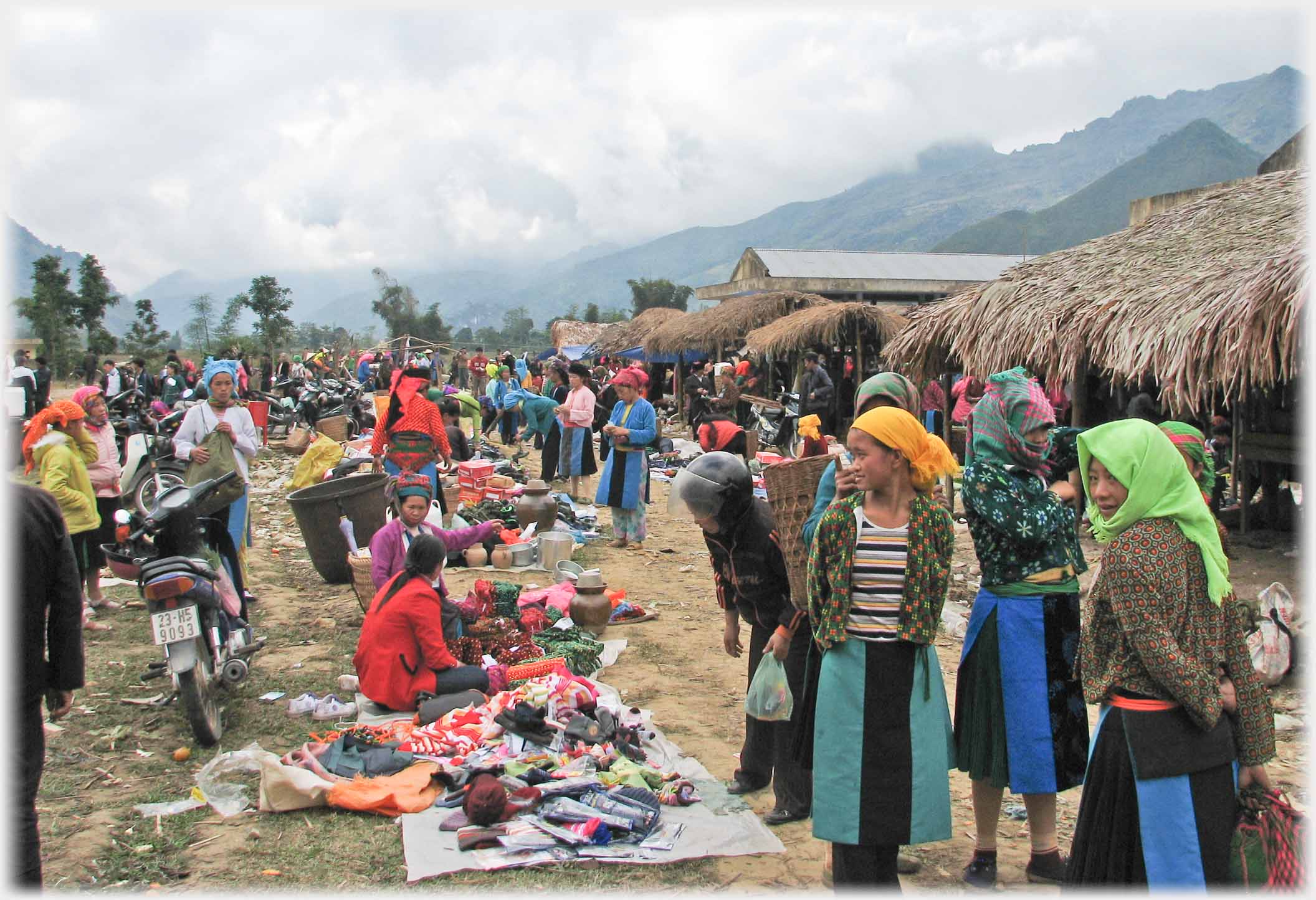 Line of sellers with goods layed out on ground in front of them, stalls in background.
