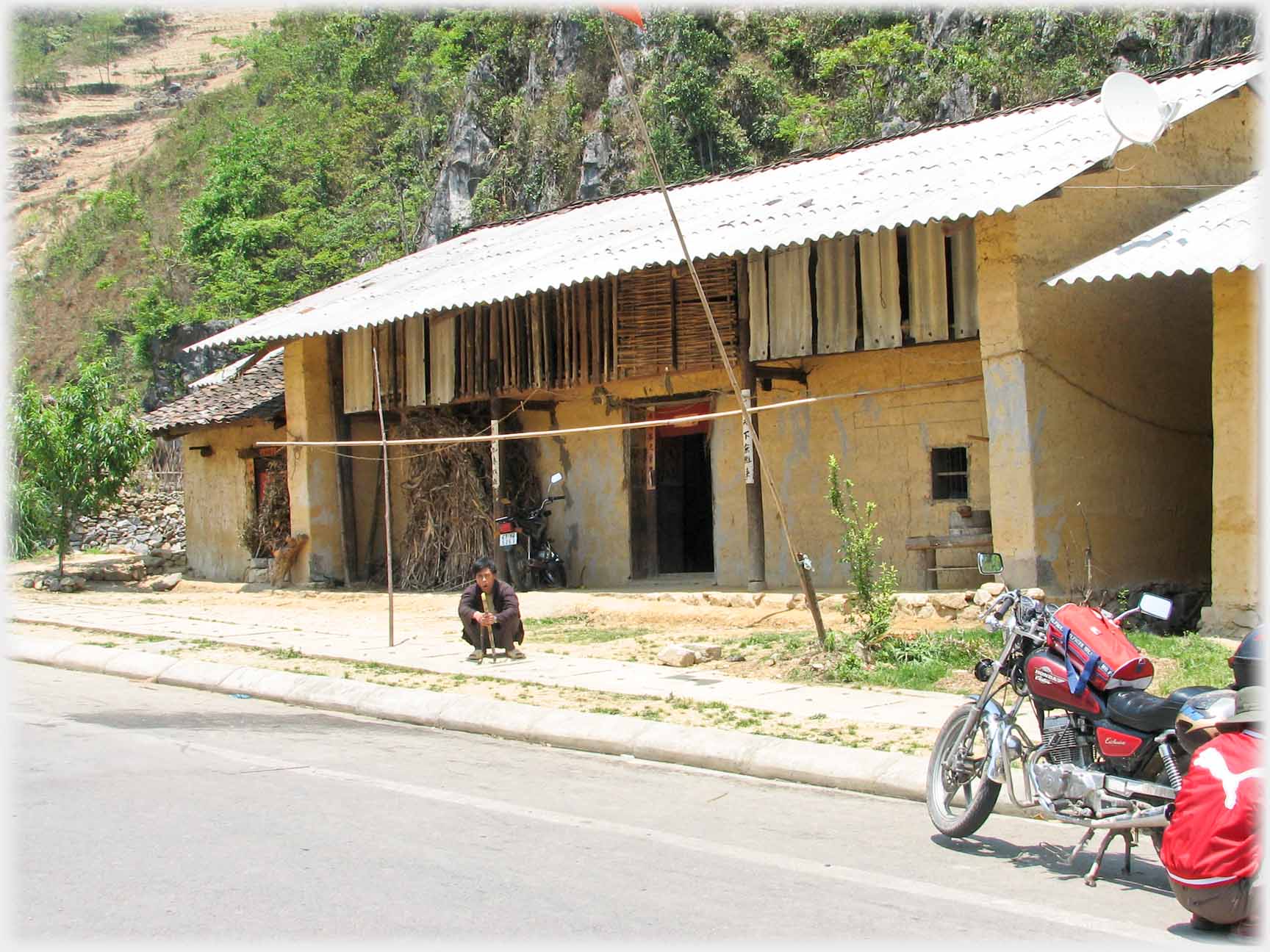 Two storied house by road with tin roof, man squatting smoking, half figure squatting by bike.