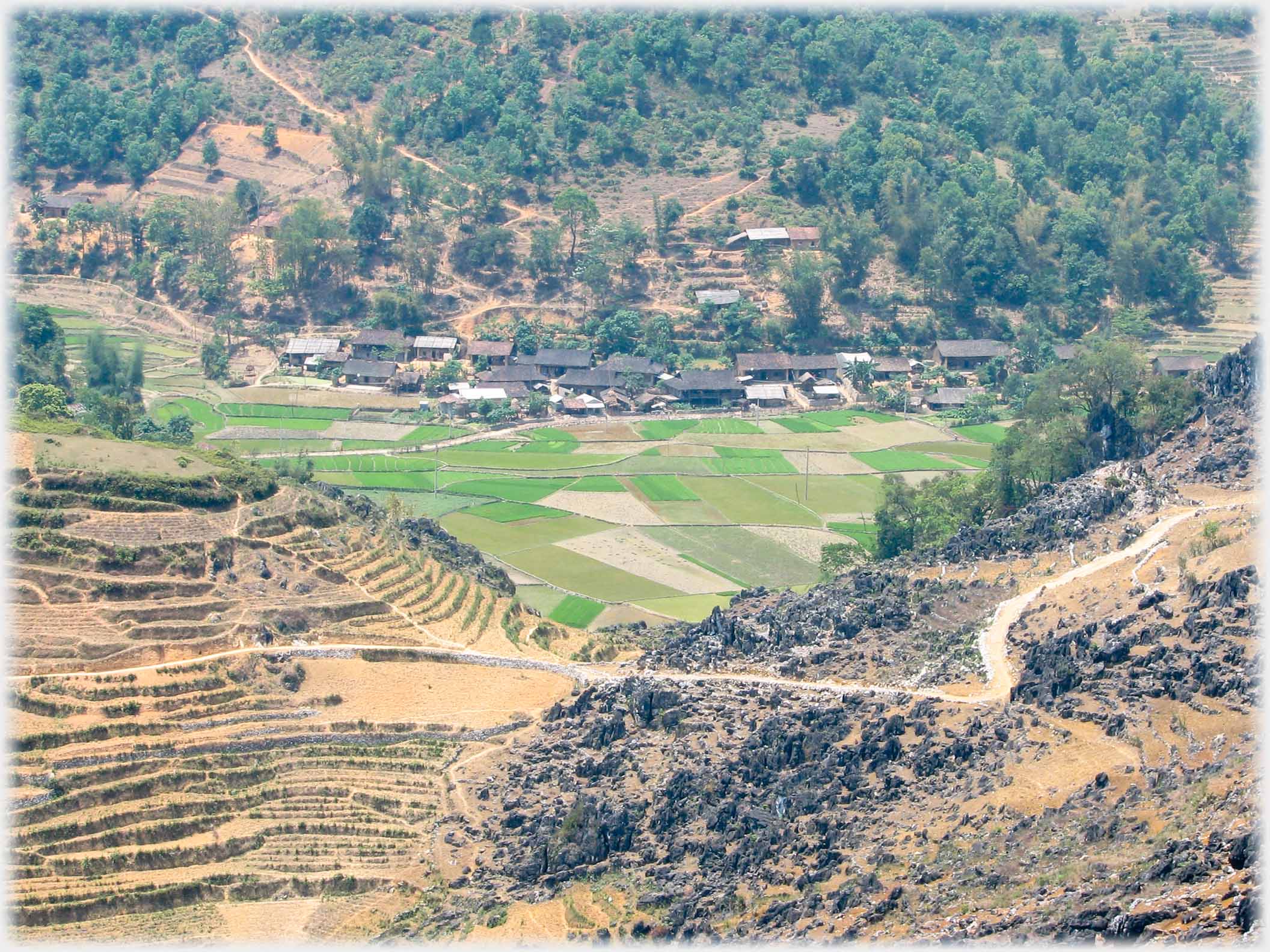 Village down in valley with patchwork of fields beside it.