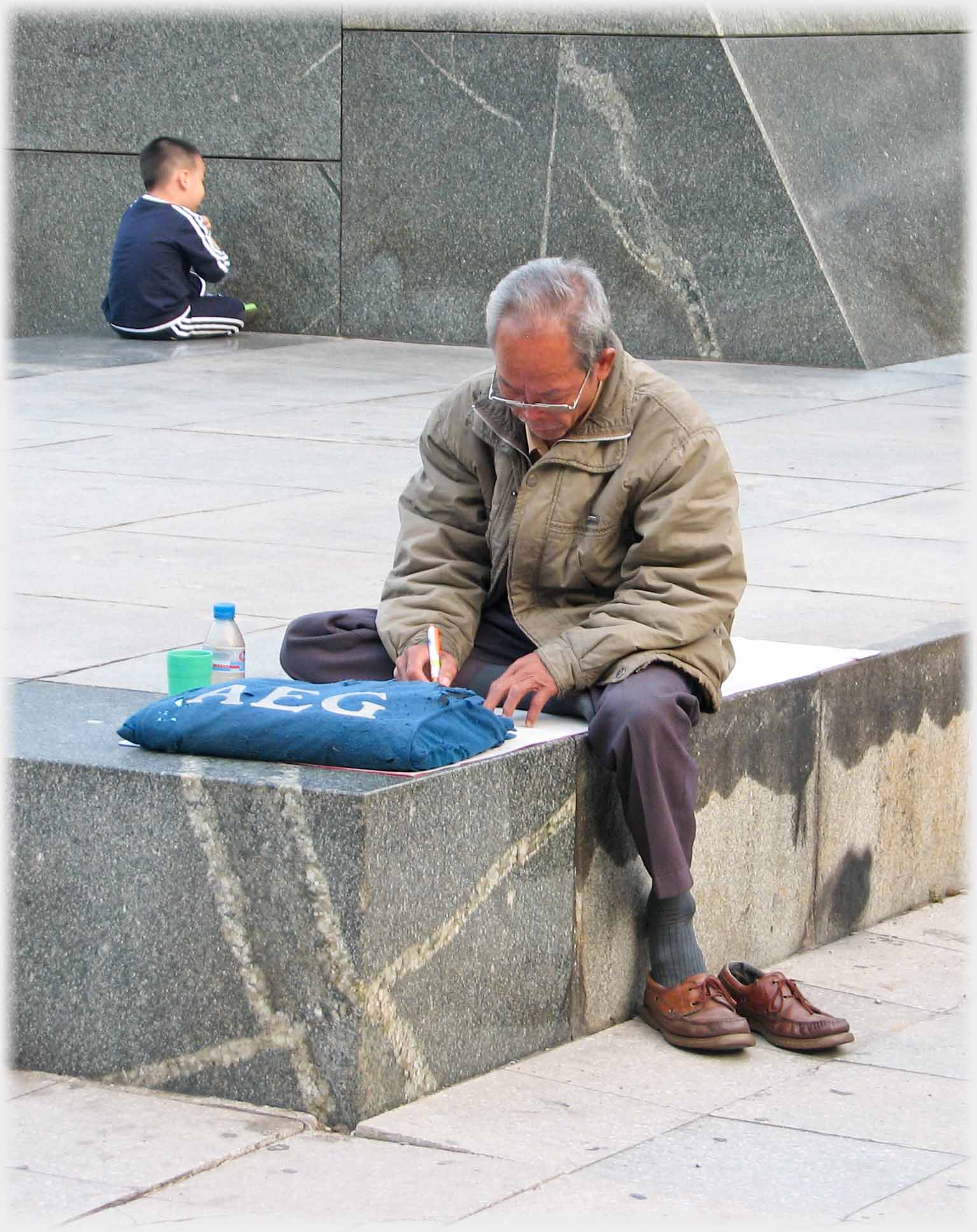 Man sitting on corner of raised area writing, one foot down.