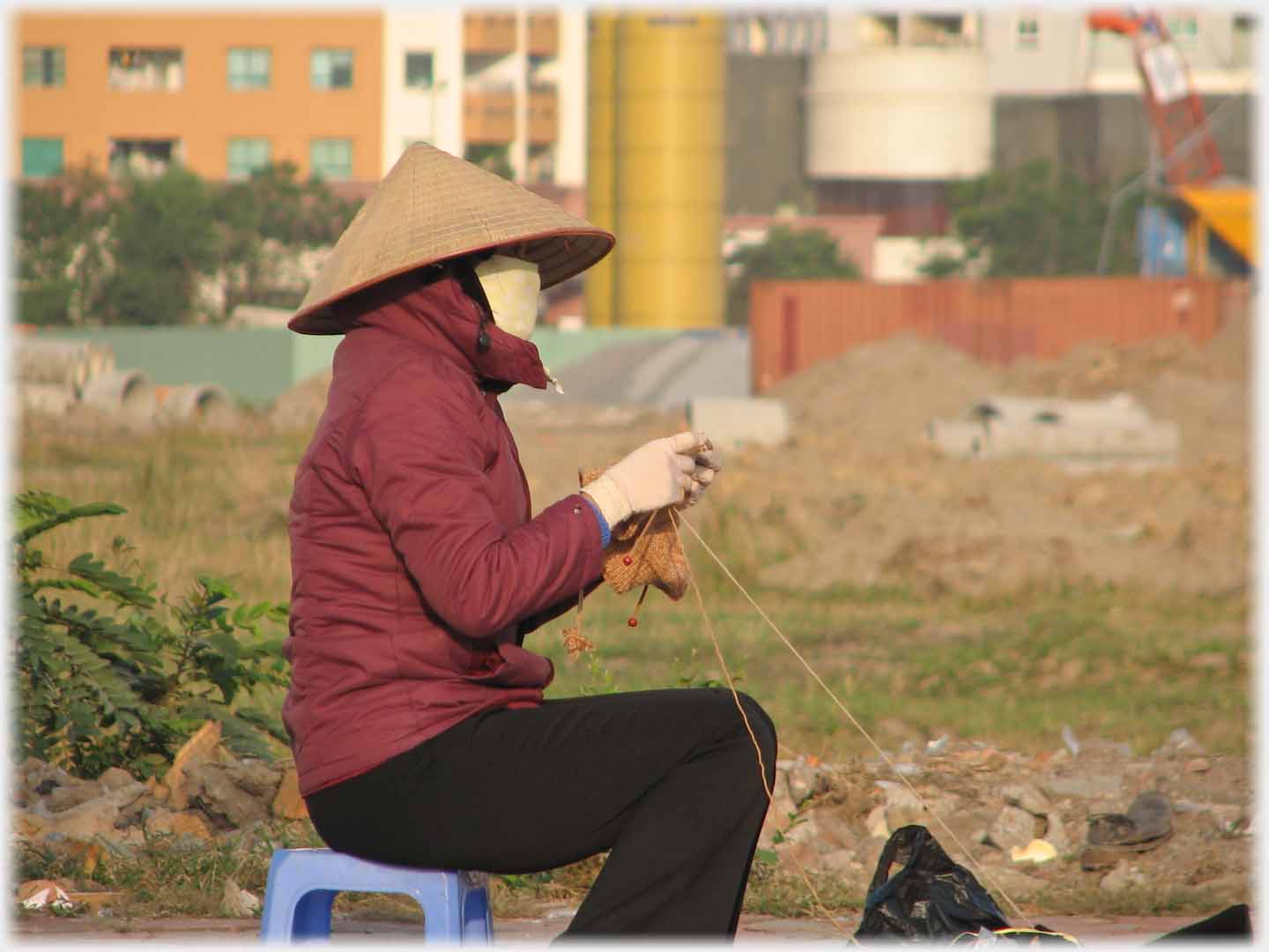 Behatted masked gloved seated woman knitting.
