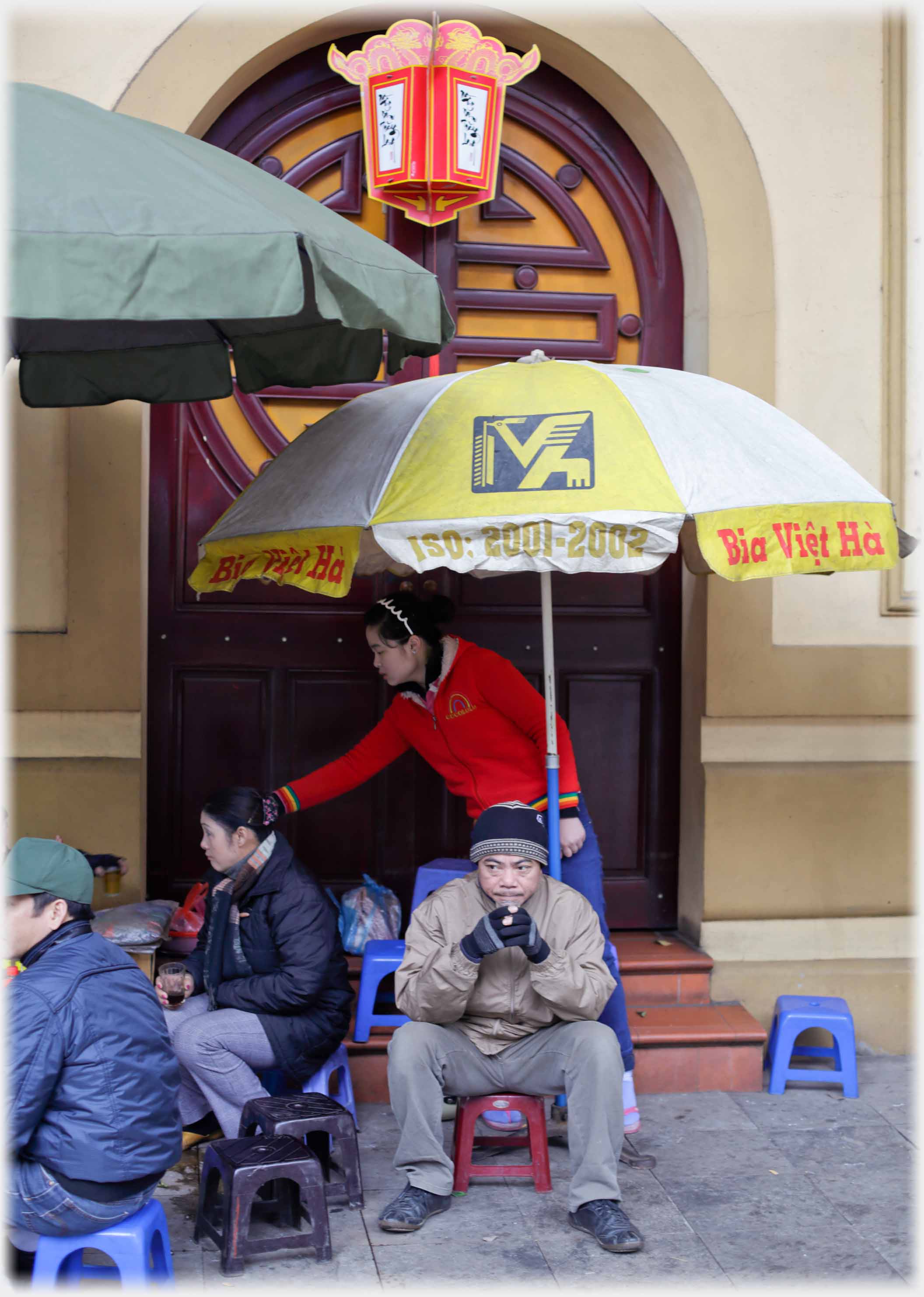 Pavement cafe with umbrella and customers looking cold, sitting on small stools.