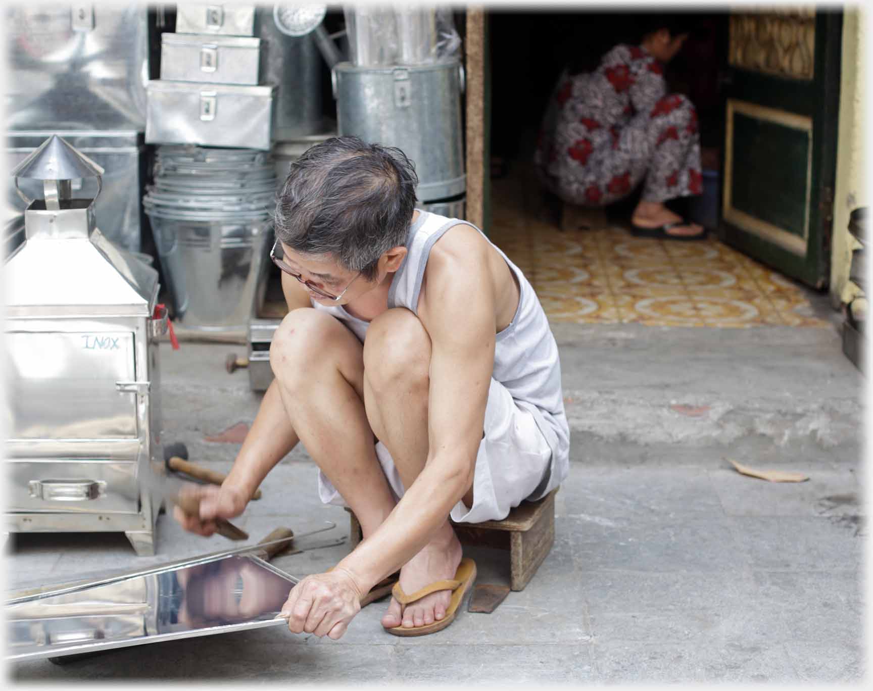 Man squatting hammering tin, woman within squatting.