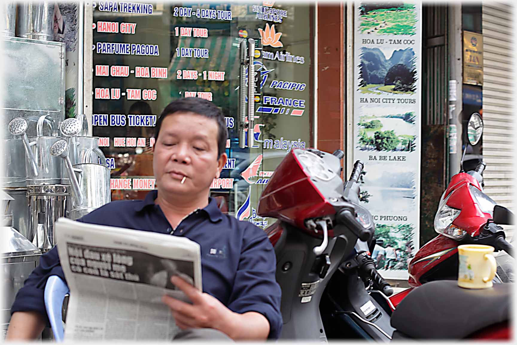 Man with contented expression toothpick in mouth reading a newspaper next to a motorbike with mug on it.
