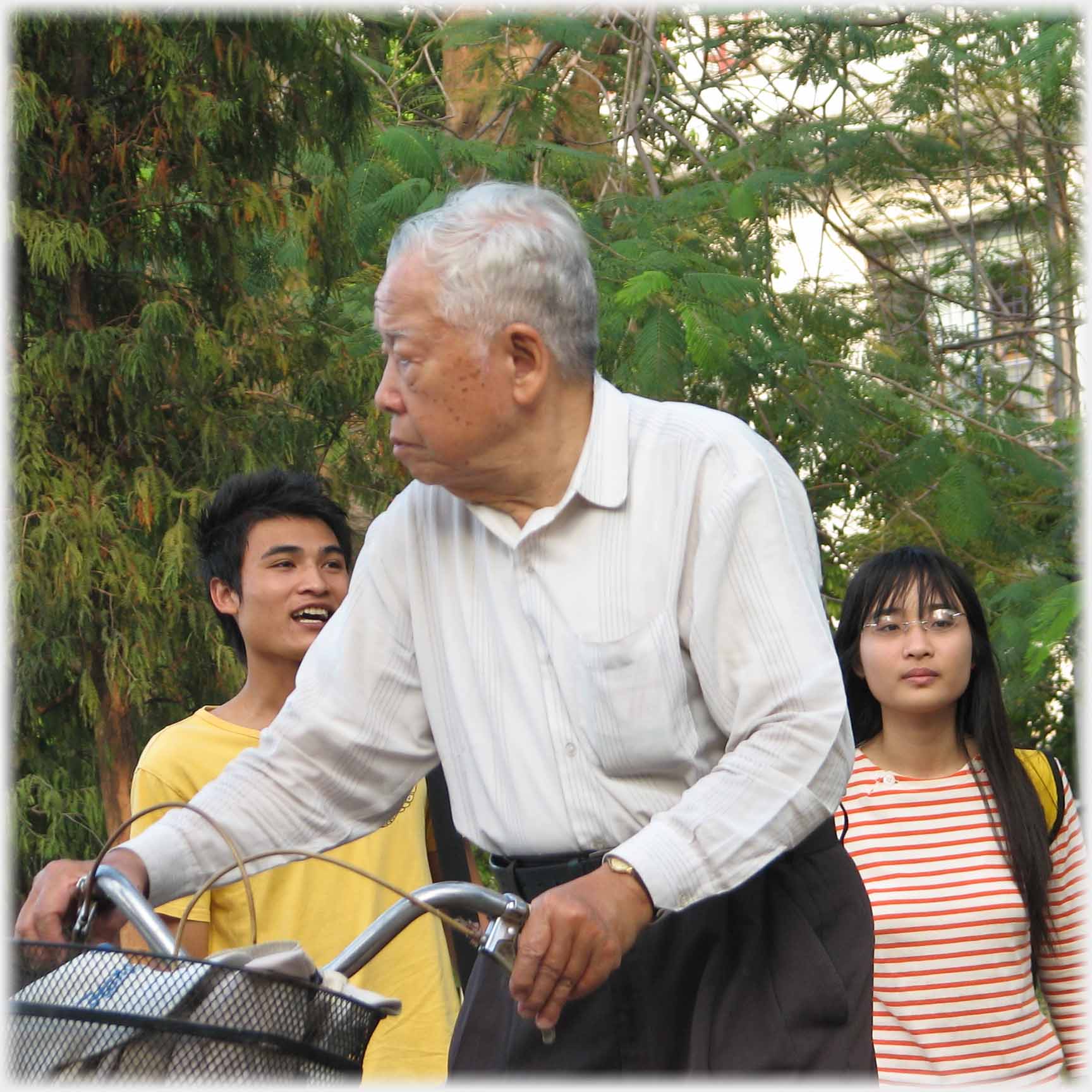 Older man wheeling bike followed by young couple.
