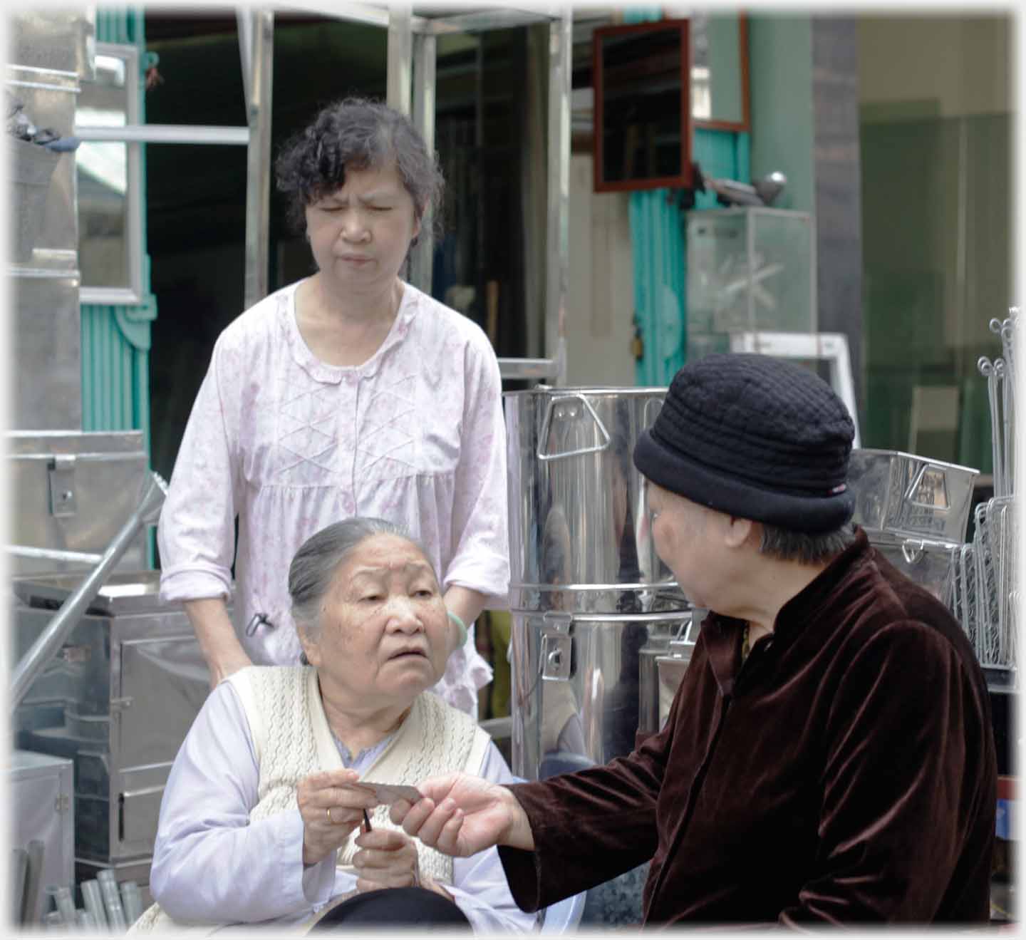 Two older women sitting together passing a small paper, younger woman standing behind.