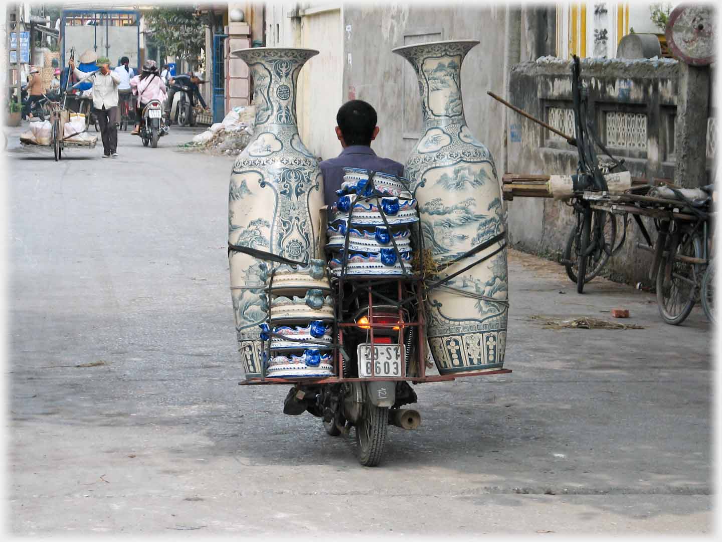 Man riding motorbike with two of the large vases rising above his head and strapped on either side of the rear pannier - looking very valnerable.