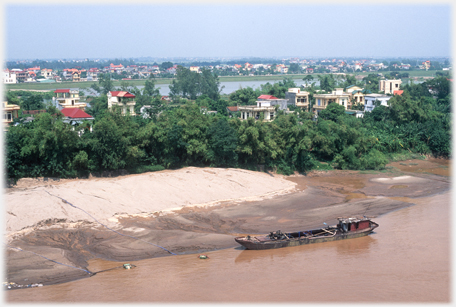 Boat on shore of river, low rise houses and trees beyond.