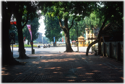 Dense shadde of trees, beyond in sun edge of yellow building and end of mausoleum.