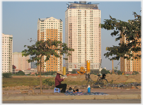 Thirty story blocks of flats with woman selling goods spread out on pavement in front.