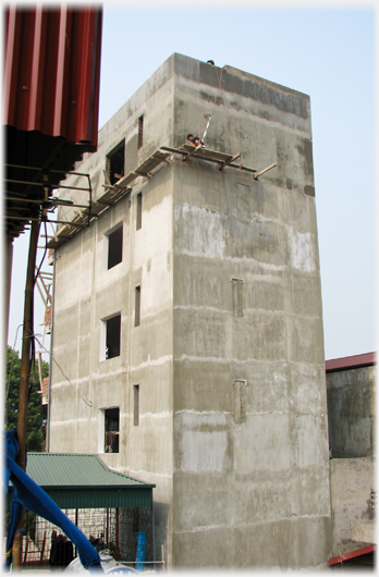 Concrete five story smooth walled block, man on small plank on wall at top level.