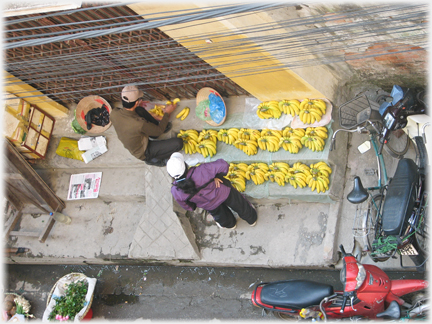 Three steps in front of a door covered in bunches of bananas, two people and motorbikes.
