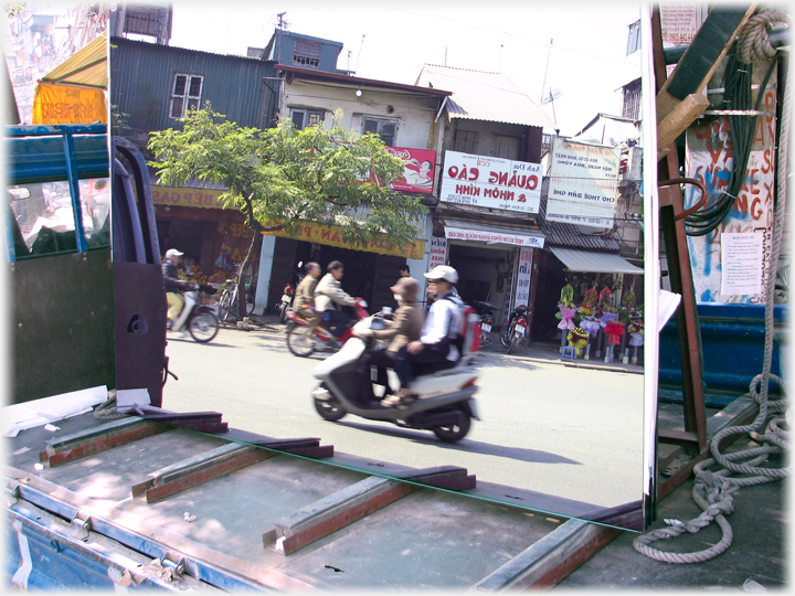 Large mirror on back of truck reflecting motorsyclists, tree and shops with reversed signs over them.