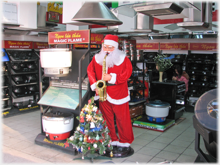 Santa manikin with sax, next to tiny artificial tree at front of gas hob shop.