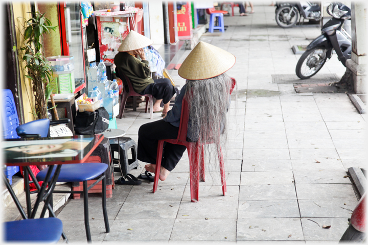 Centre of picture on pavement, woman's back on plastic chair, grey hair to the ground, conical hat down to chair back, another woman beyond.