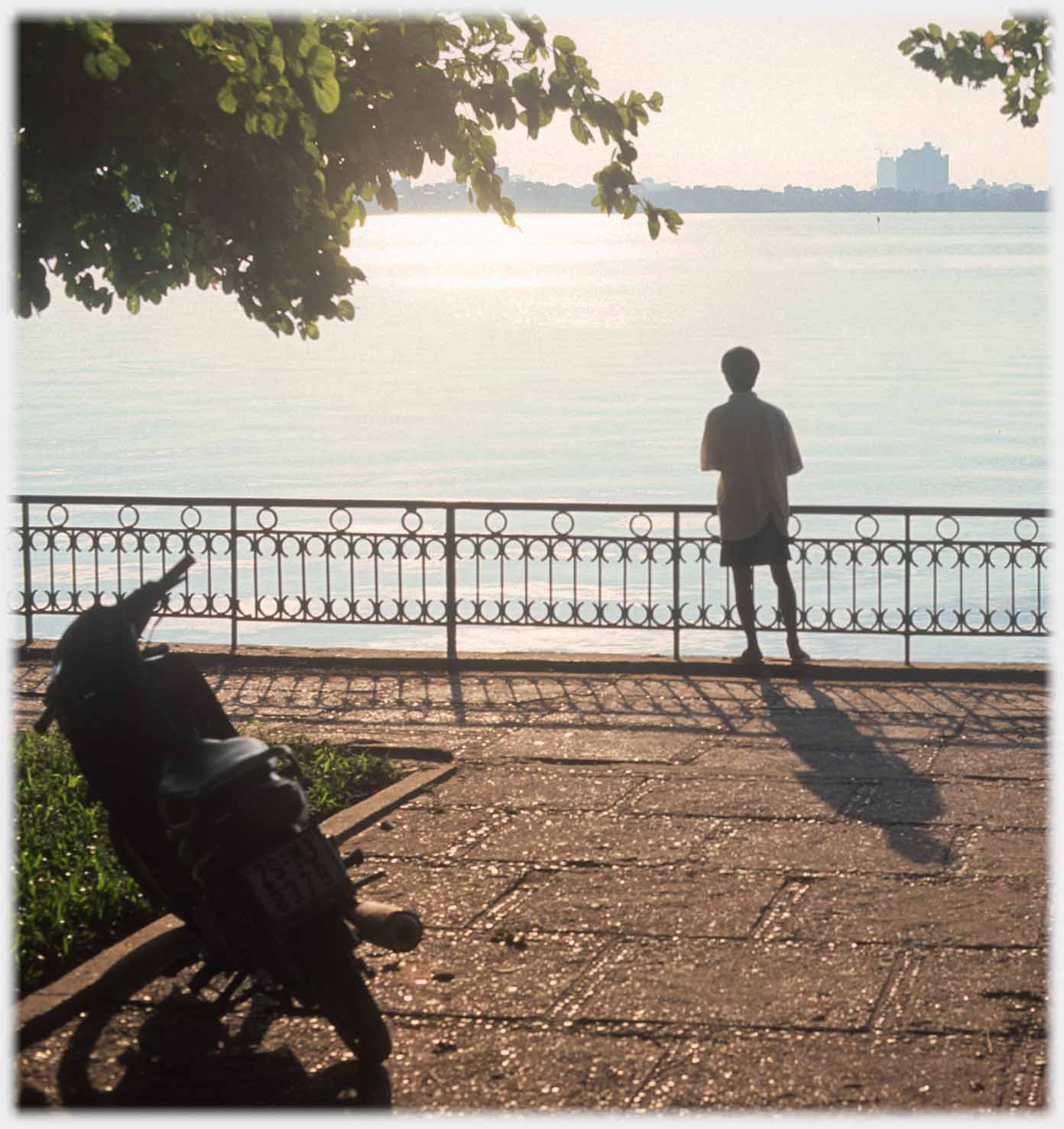 Man with long shadow by lake railings.