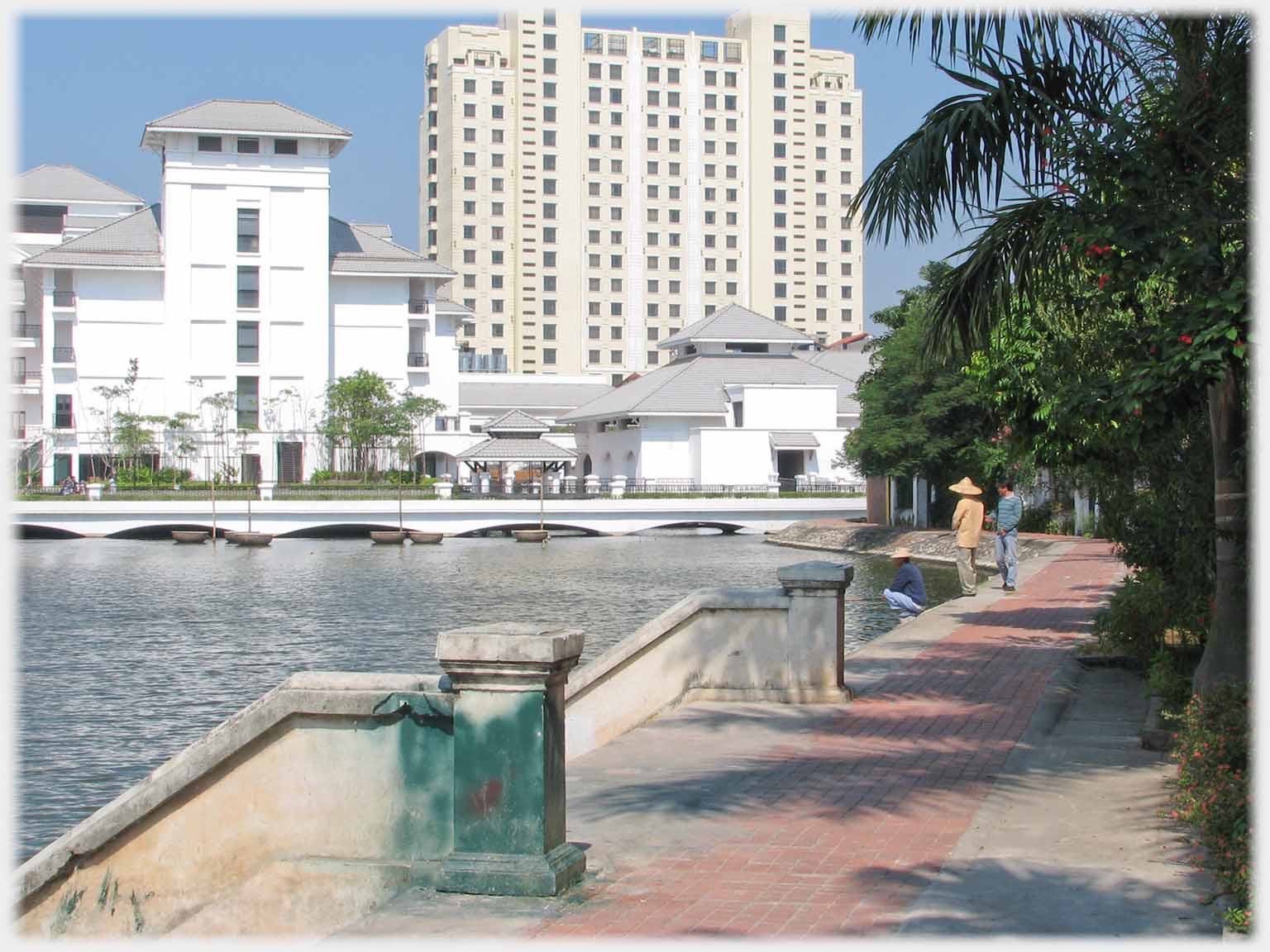 Skyscraper above lower white buildings, three men at water's edge nearer.