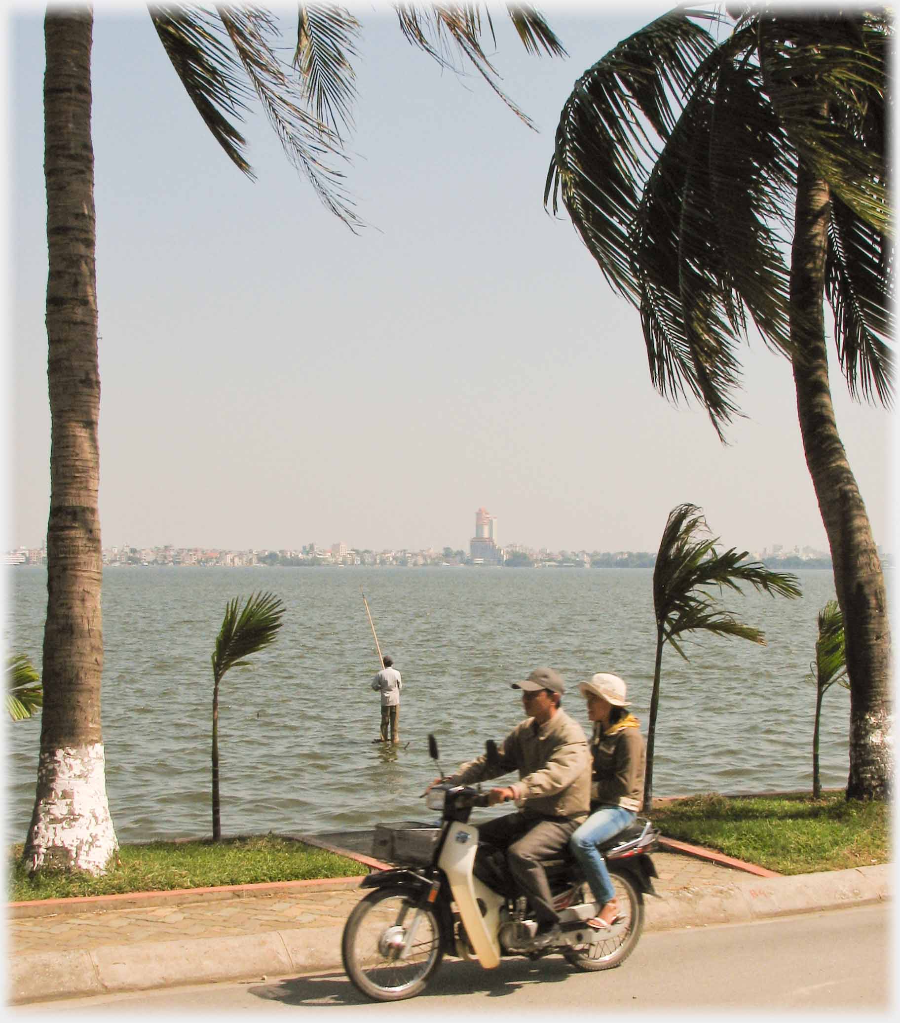 Couple on motorbike passing trees and fisherman.