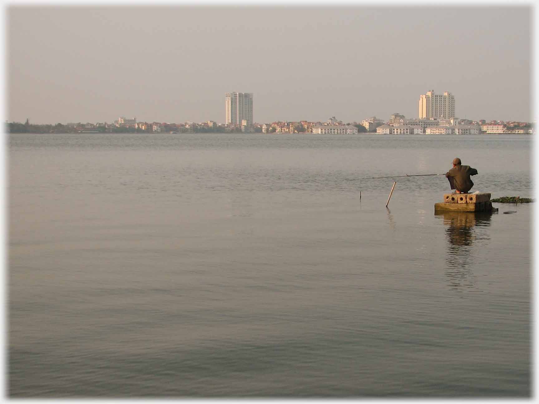 Man Man squatting on blocks in lake, far horizon of city - continued in next picture.