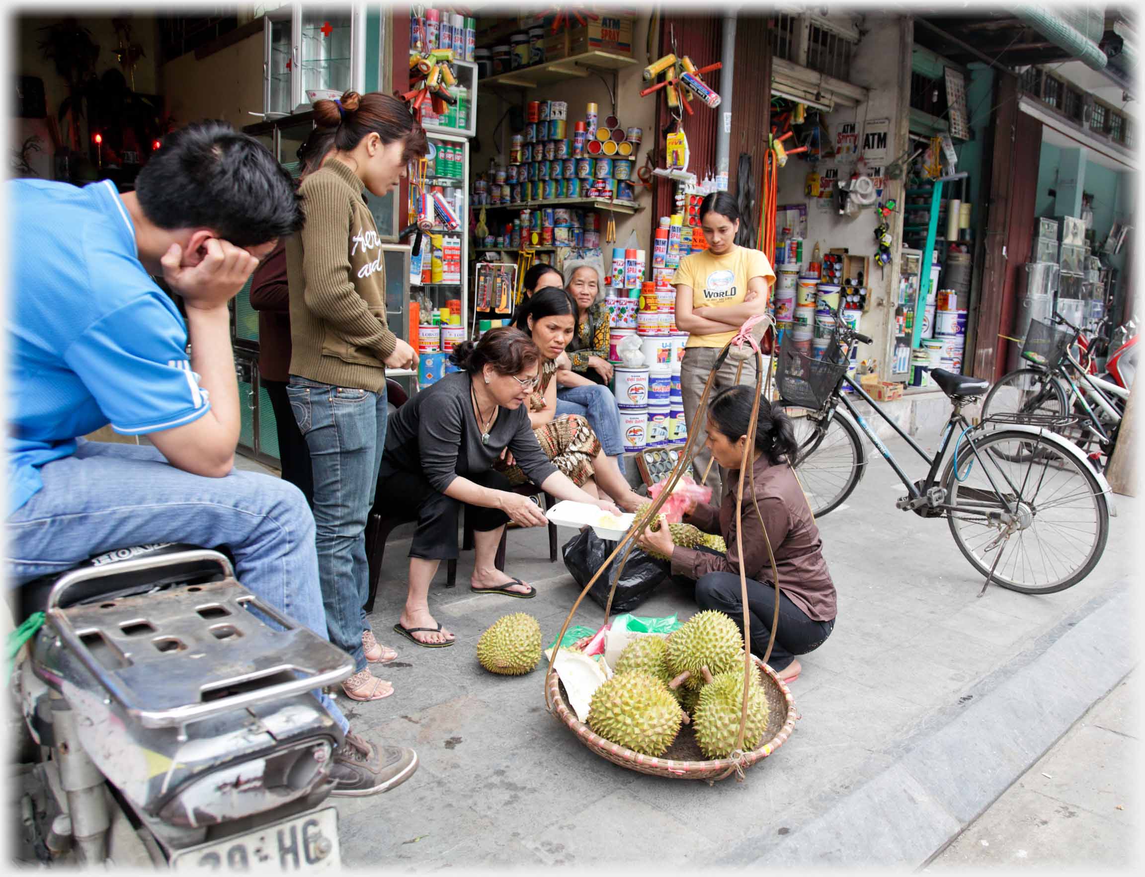 Six women and a man on a motorbike watching a woman cut up a durian.