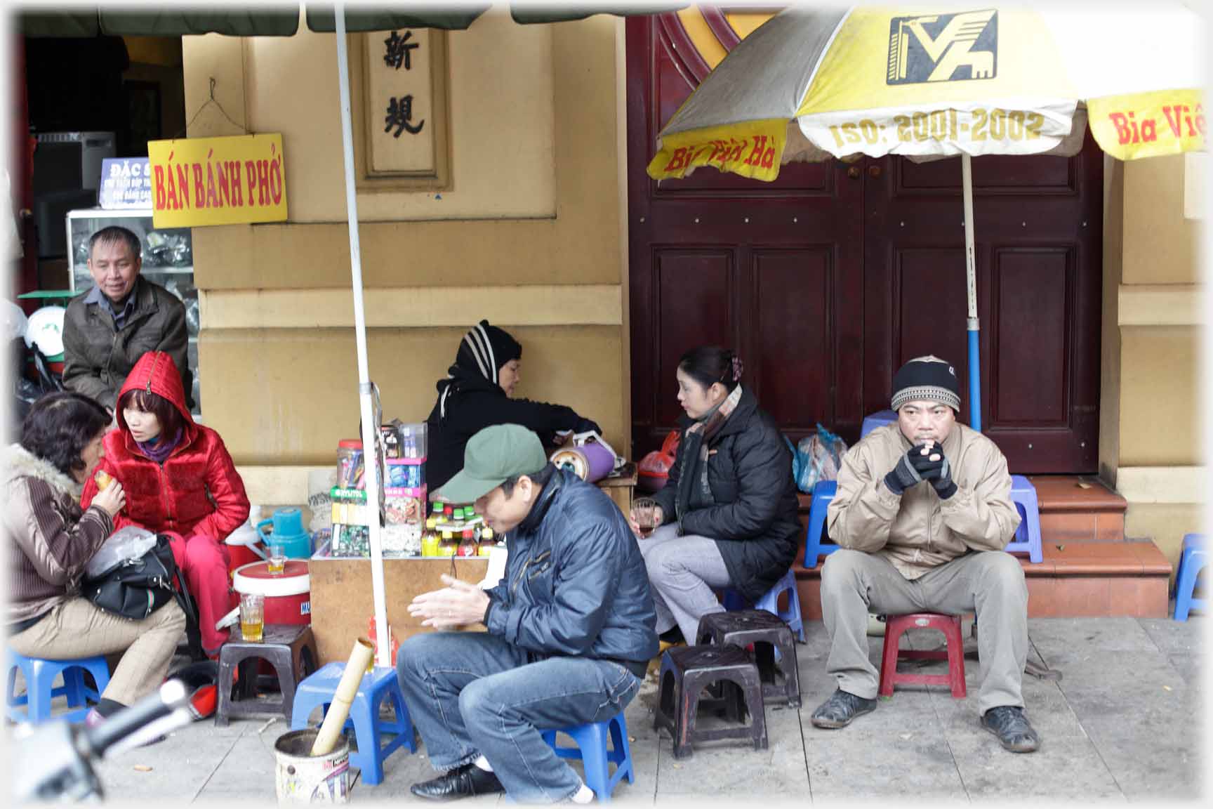 Group of people on low stools in winter clothes by woman pouring tea.