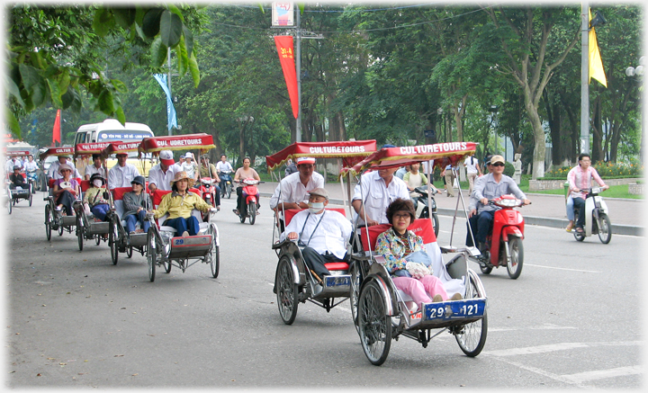 Line of rickshaws moving down wide street with motorbikes.