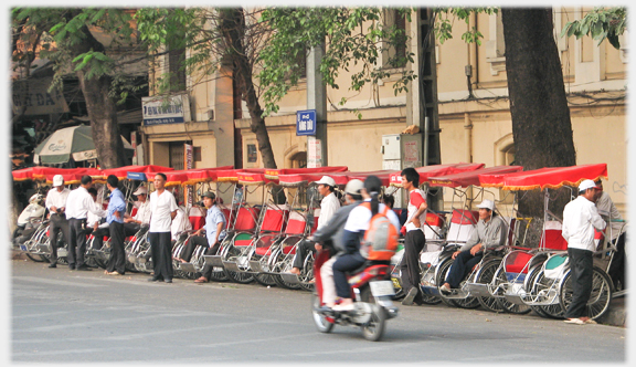 Line of parked rickshaws under trees at side of road.